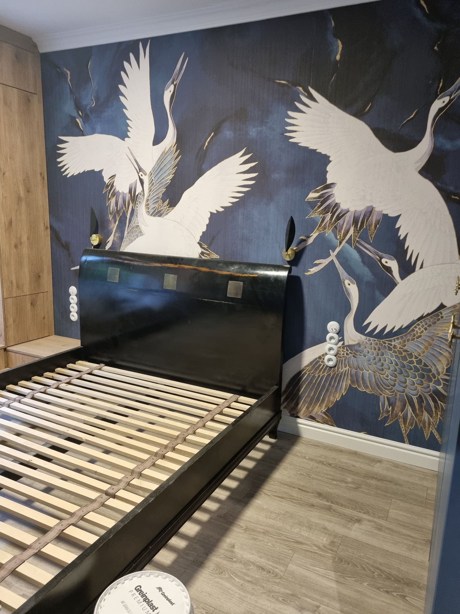 Ściana z motywem żurawi za czarną ramą łóżka z drewnianym stelażem, widoczna puszka z farbą na podłodze.