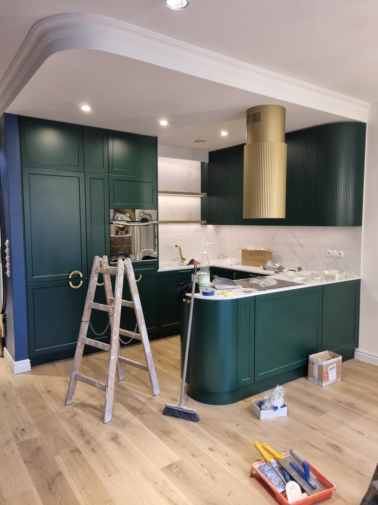 Nowoczesna kuchnia w odcieniach zieleni z drewnianą podłogą w trakcie prac wykończeniowych, widoczna drabina malarska i akcesoria malarskie, złoty okap nad wyspą kuchenną.