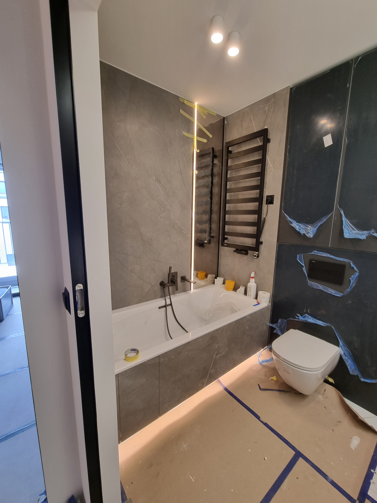 Wnętrze łazienki w trakcie remontu: biała wanna obudowana szarą płytką imitującą marmur, podwieszana toaleta, grzejnik drabinkowy, zabezpieczone folią płytki na ścianach, podłoga wyłożona kartonem.