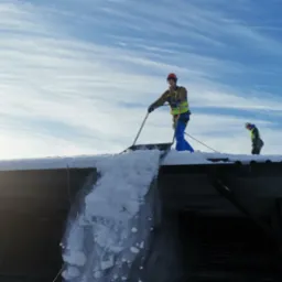 Dwóch pracowników w kaskach i kamizelkach odblaskowych odśnieża dach budynku za pomocą łopat, zrzucając śnieg na dół na tle błękitnego nieba z chmurami.