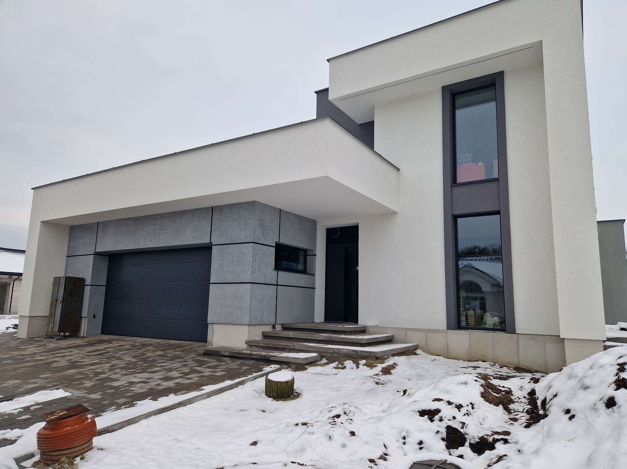 Nowoczesny dom jednorodzinny z płaskim dachem, wykończony białym i szarym tynkiem elewacyjnym, z widocznymi schodami wejściowymi i bramą garażową, otoczony resztkami śniegu.