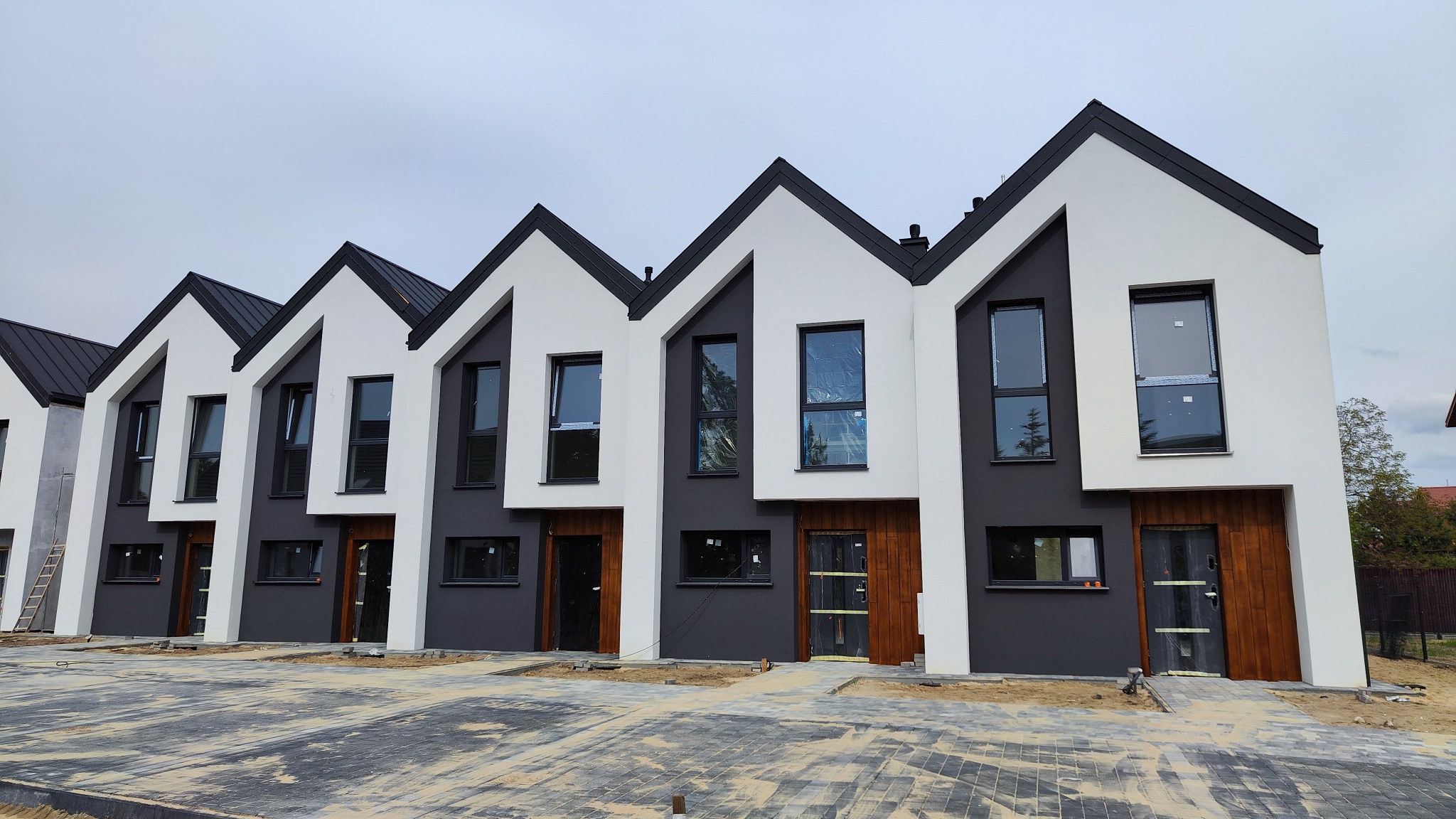 Szereg identycznych, nowoczesnych domów jednorodzinnych w trakcie budowy, z pomalowanymi elewacjami na biało i szaro, czarnymi dachami i drewnianymi drzwiami wejściowymi.