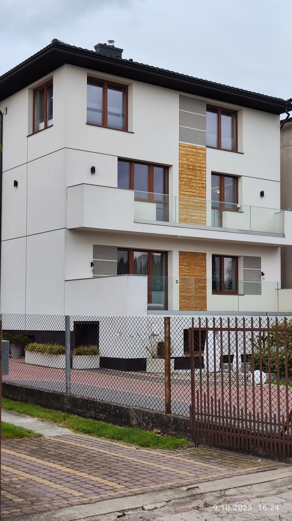 Nowoczesny, dwupiętrowy dom z białymi ścianami, drewnianymi i szarymi akcentami elewacyjnymi oraz przeszklonymi balkonami, widziany zza metalowego ogrodzenia