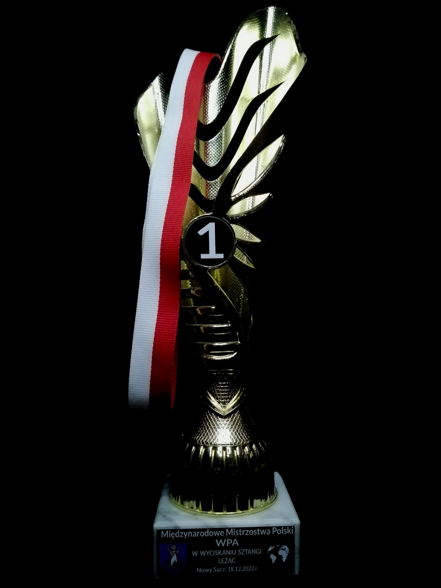 Złoty puchar z numerem 1 i biało-czerwoną wstążką, na marmurowej podstawie z napisem 'Międzynarodowe Mistrzostwa Polski WPA', na czarnym tle.
