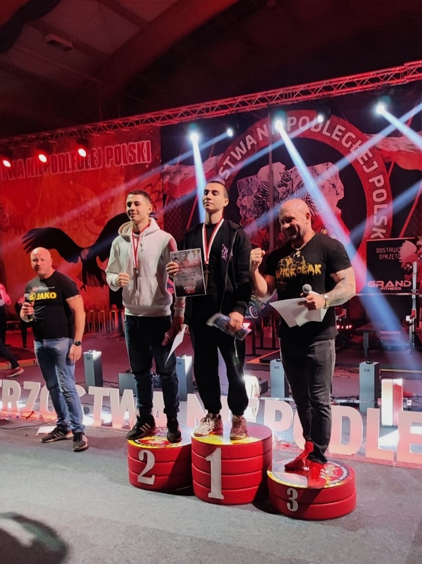 Trzech mężczyzn na podium po zawodach sportowych, jeden z mikrofonem wręcza nagrody, w tle baner z napisem.