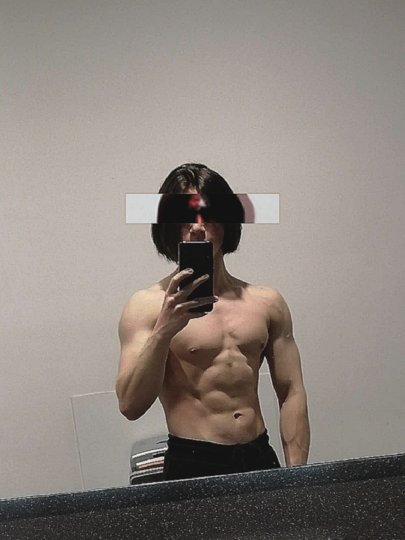 Mężczyzna z zamazaną twarzą robi selfie, prezentując wyrzeźbioną sylwetkę, widoczne mięśnie brzucha i ramion, czarne spodnie, odbicie w lustrze.