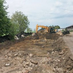 Usługi remontowo wykonczeniowe - Żółta koparka Hyundai na placu budowy, przygotowuje teren pod budowę. Stosy opon i drewna w tle, obok budynek gospodarczy. Pochmurne niebo.