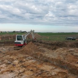 Usługi remontowo wykonczeniowe - Biała koparka w wykopie na placu budowy, w tle zielone pole i pochmurne niebo. Widoczne drewniane paliki wyznaczające granice. Na ziemi leży miarka.