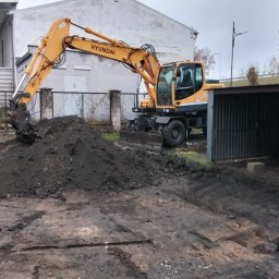 Usługi remontowo wykonczeniowe - Żółta koparka Hyundai na placu budowy, z hałdą ziemi przed łyżką. W tle budynek i ogrodzenie z siatki.