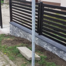 Usługi remontowo wykonczeniowe - Nowoczesne ogrodzenie panelowe z poziomymi przęsłami w kolorze czarnym, osadzone na podmurówce z szarej kostki. Widok z poziomu gruntu, trawa i kostka brukowa w tle.