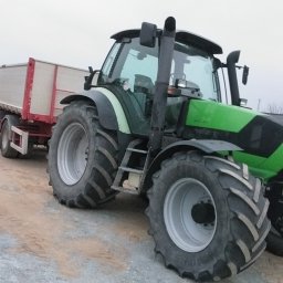 Usługi remontowo wykonczeniowe - Zielony traktor z przyczepą na placu budowy, w tle widoczna koparka. Ujęcie z boku, podkreślające masywność maszyny i jej gotowość do pracy.