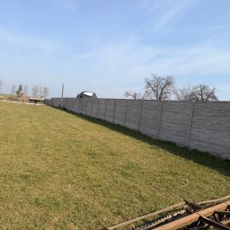 Usługi remontowo wykonczeniowe - Długi, szary, betonowy płot panelowy na zielonym trawniku pod błękitnym niebem. W rogu fragment brony talerzowej. Widok na posesję.