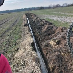 Usługi remontowo wykonczeniowe - Wykopany rów melioracyjny na polu, wypełniony wodą. Widoczne ślady opon i resztki roślinności. W tle las i błękitne niebo.