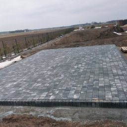 Usługi remontowo wykonczeniowe - Ułożona, szara kostka brukowa na przygotowanym podłożu. Widoczne obrzeża, w tle krajobraz z drzewami i polem. Częściowo leży śnieg.