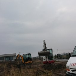 Usługi remontowo wykonczeniowe - Dwie koparki na placu budowy, jedna z podniesioną łyżką, w tle budynek i grupa osób, z prawej strony kadru fragment białego samochodu dostawczego.