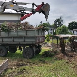 Usługi remontowo wykonczeniowe - Koparka z chwytakiem załadowuje gałęzie do przyczepy na tle zabudowań. Widoczne ślady pracy w terenie.