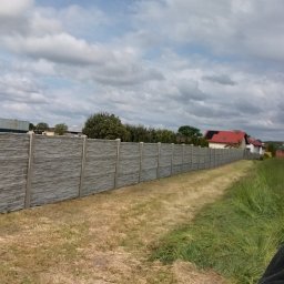 Usługi remontowo wykonczeniowe - Długi, szary, betonowy płot panelowy oddzielający posesje, widziany z perspektywy trawnika. W tle domy i pochmurne niebo.