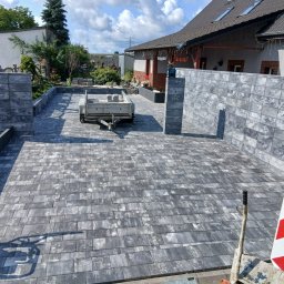 Usługi remontowo wykonczeniowe - Nowo ułożona nawierzchnia z ciemnoszarych, prostokątnych elementów betonowych, otoczona pasującymi murkami z bloczków. W tle dom, mała przyczepa i zagęszczarka.
