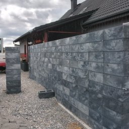 Usługi remontowo wykonczeniowe - Nowoczesny mur z szarych, fakturowanych bloczków betonowych, częściowo połączony z ceglaną ścianą. Obok domu z ciemnym dachem, na żwirowym podłożu, widoczna mini-koparka.