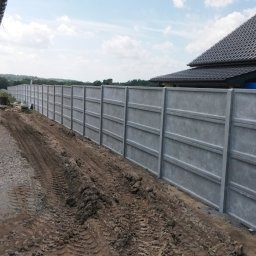 Usługi remontowo wykonczeniowe - Długie, szare ogrodzenie z prefabrykowanych paneli betonowych z poziomymi przetłoczeniami, biegnące wzdłuż boku budynku. Na pierwszym planie błotnista ziemia ze śladami opon, w tle zielony krajobraz.
