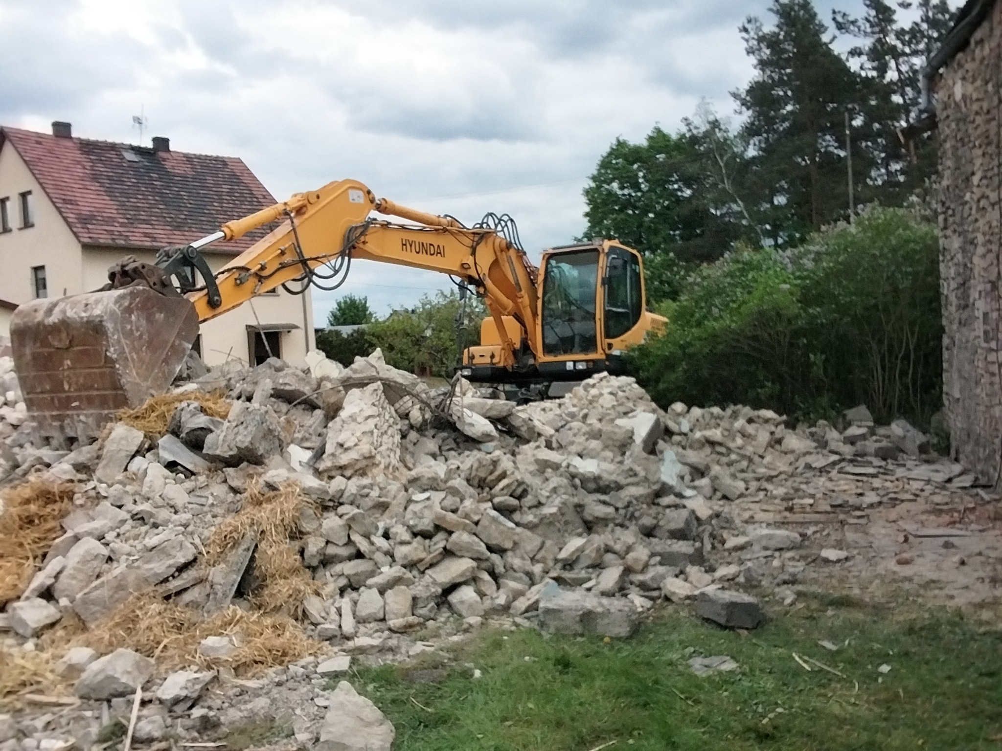 Żółta koparka Hyundai na gruzowisku po rozbiórce w Strzelcach Opolskich. Widoczny fragment budynku i drzewa w tle. Prace wyburzeniowe w toku.