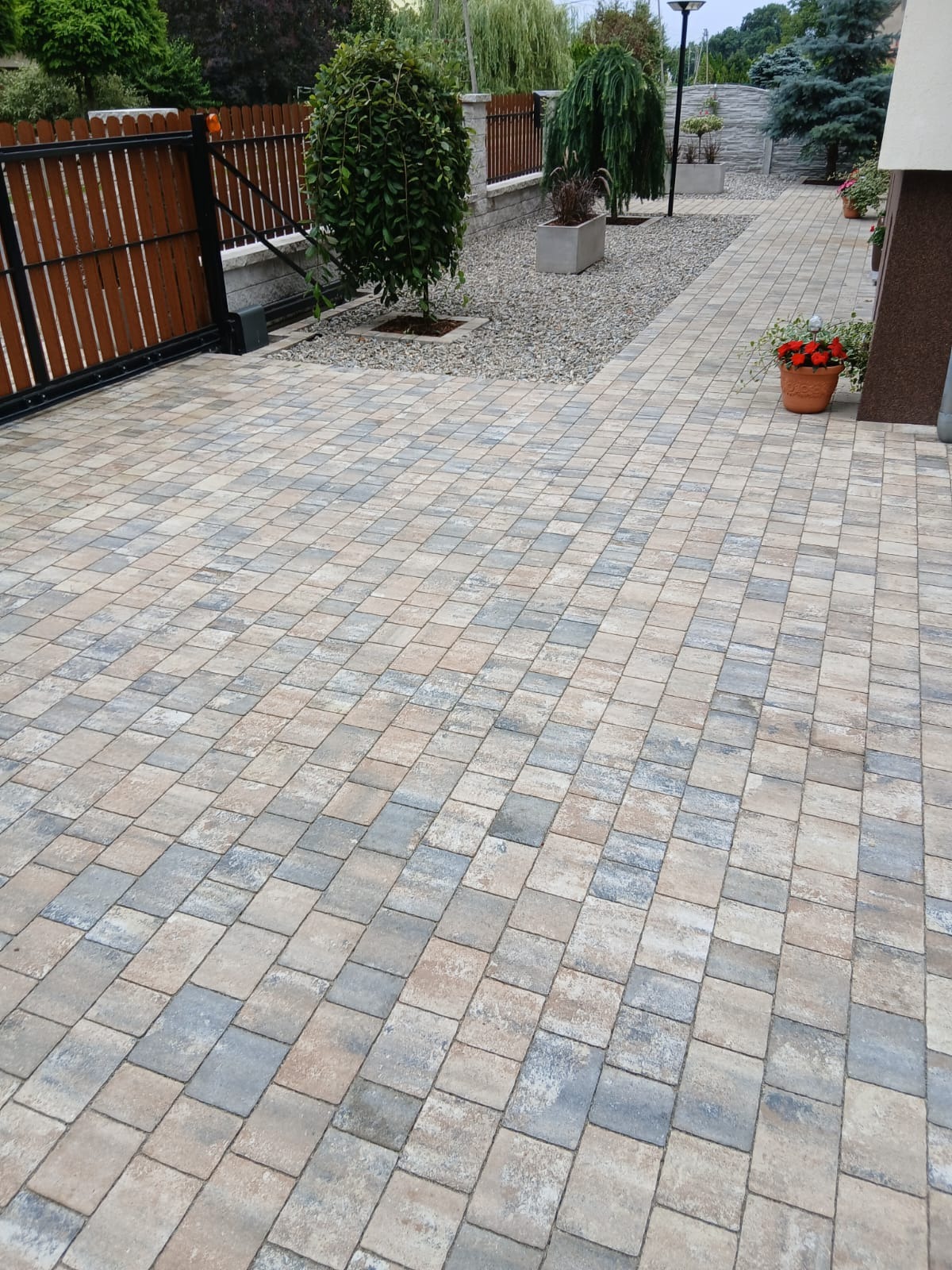 Ułożona kostka brukowa w odcieniach beżu i szarości tworzy wzór na podjeździe. Widoczny fragment ogrodzenia panelowego i roślinność w tle. Czerwone kwiaty w donicy dodają koloru.