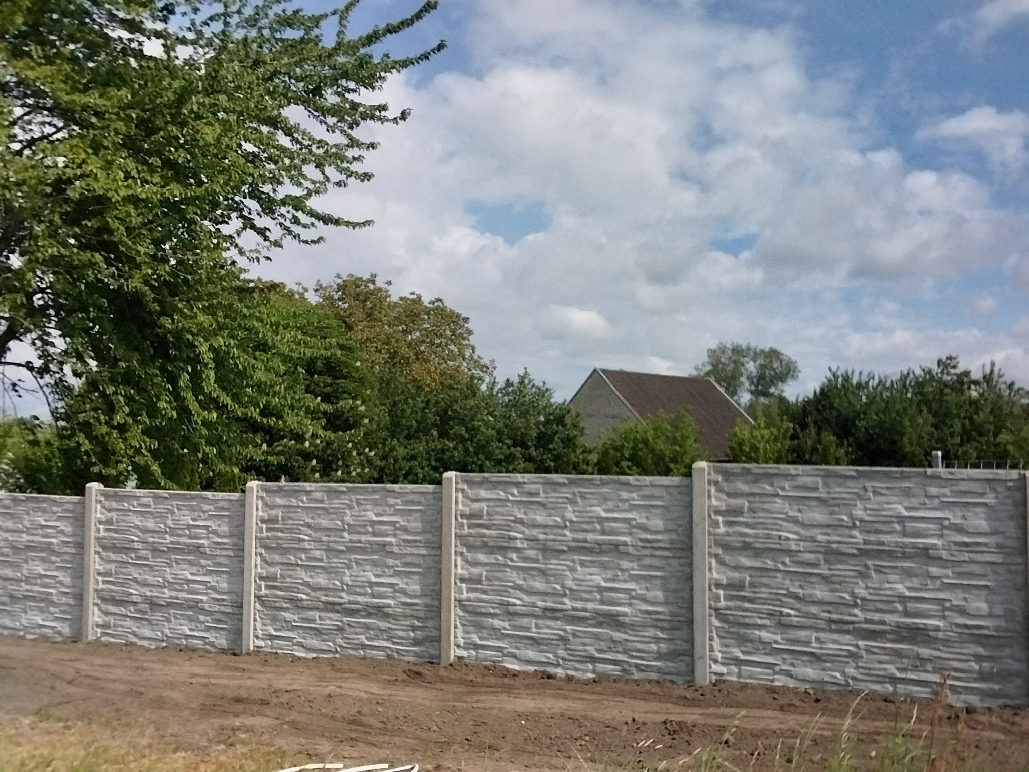 Gotowy, szary, betonowy płot panelowy z fakturą kamienia, oddzielający teren zielony. Widok na drzewa i fragment dachu domu w tle.