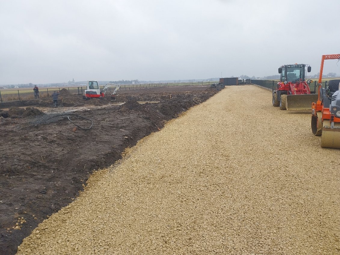 Przygotowanie terenu pod budowę: wyrównany teren z kruszywem, mini koparka i walec w tle.  Pracownicy w oddali. Pochmurna pogoda.