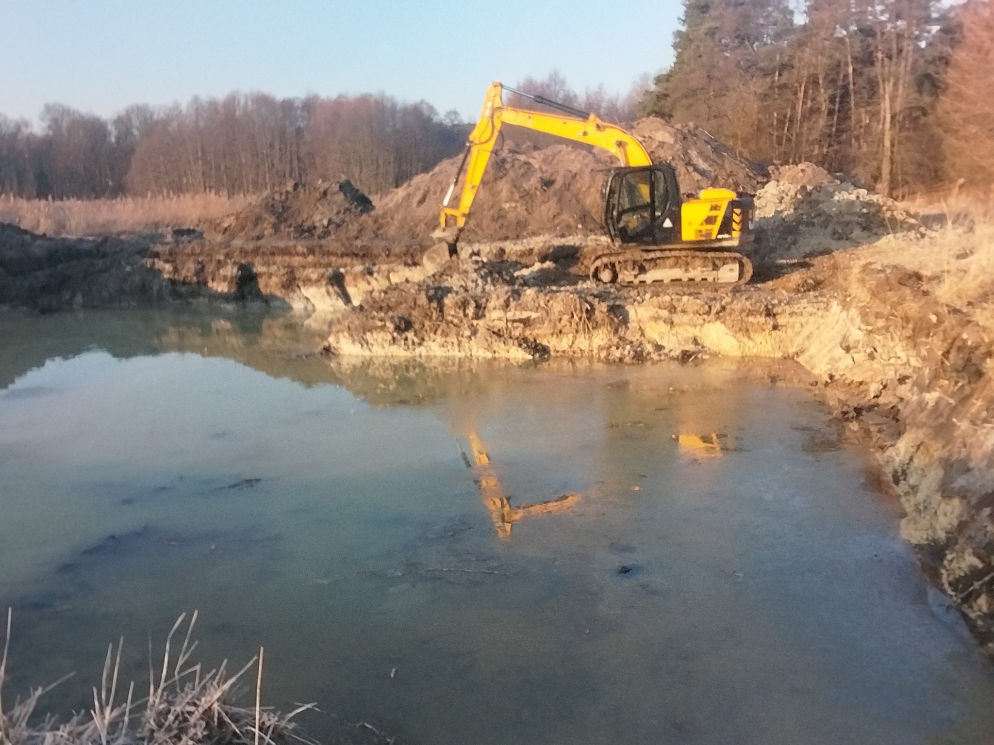 Żółta koparka na brzegu wykopu wypełnionego wodą, odbicie maszyny w tafli wody. W tle drzewa i hałdy ziemi. Wczesny etap prac ziemnych.