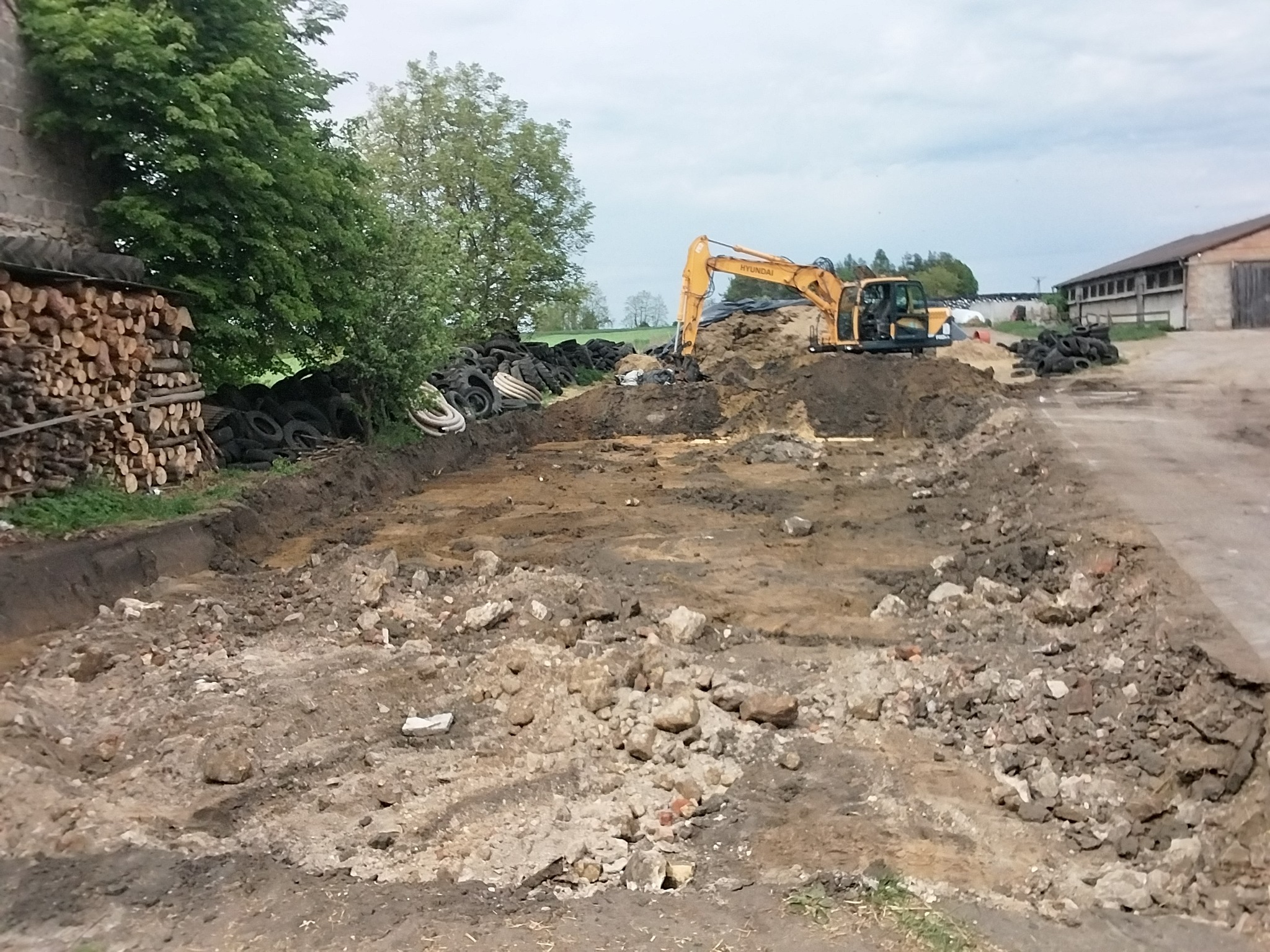 Żółta koparka Hyundai na placu budowy z wykopem, stertą opon i stosem drewna w tle. Teren przygotowany pod inwestycję, widoczne ślady pracy maszyny.