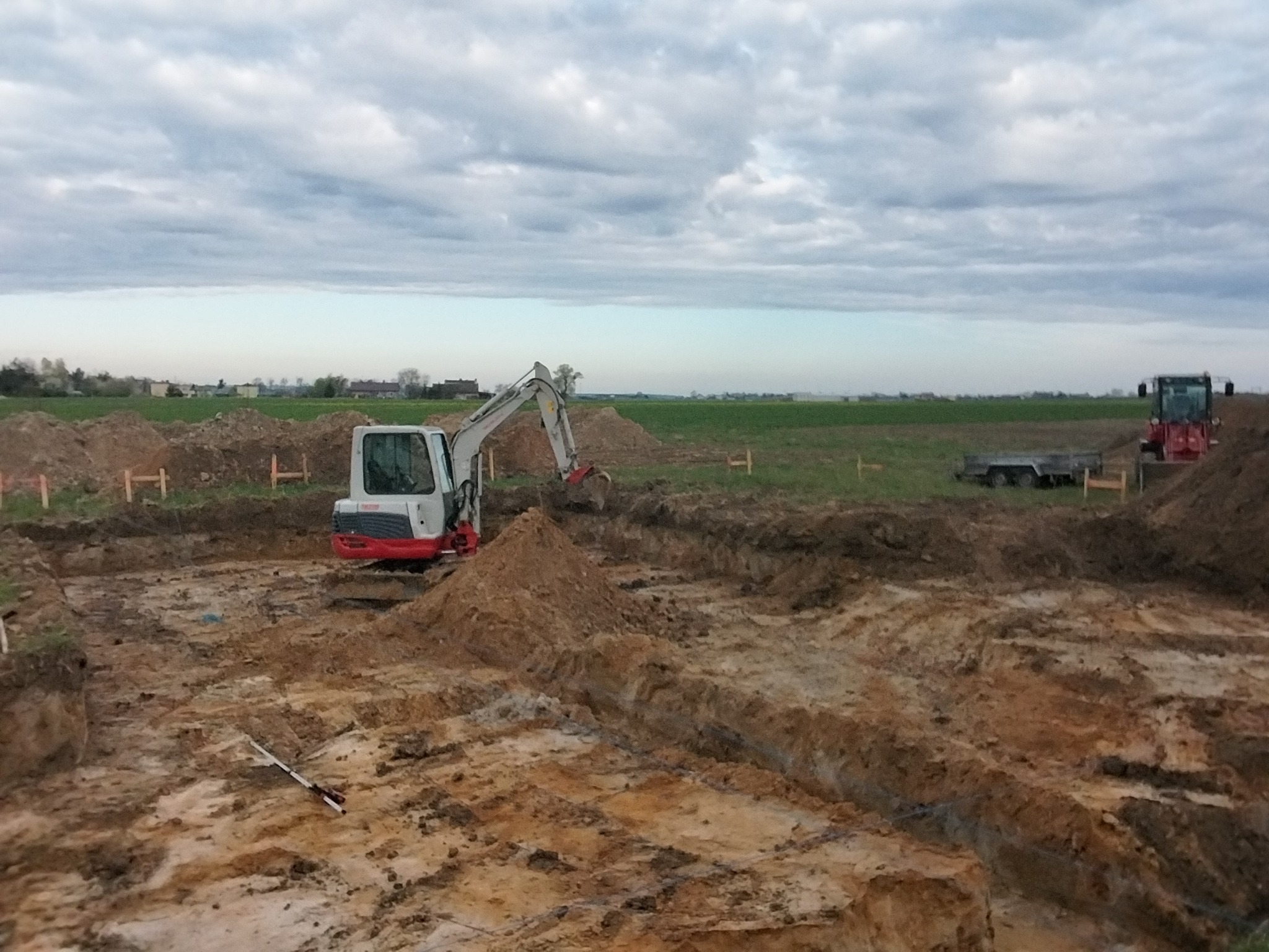 Biała koparka w wykopie na placu budowy, w tle zielone pole i pochmurne niebo. Widoczne drewniane paliki wyznaczające granice. Na ziemi leży miarka.