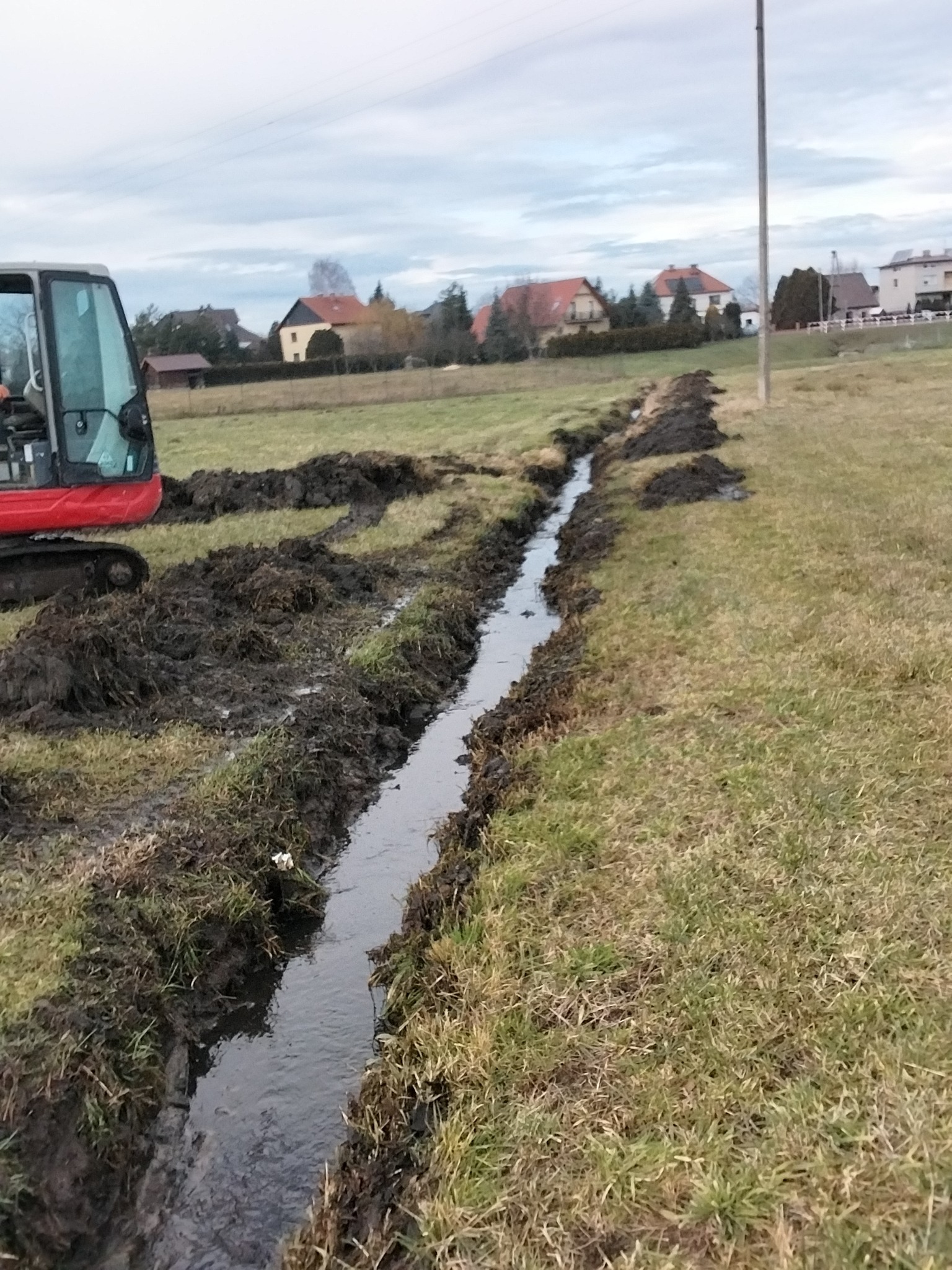 Wykonanie rowu odwadniającego na łące, z użyciem czerwonej mini-koparki, widok na wykopany rów wypełniony wodą, w tle zabudowania wiejskie pod pochmurnym niebem.