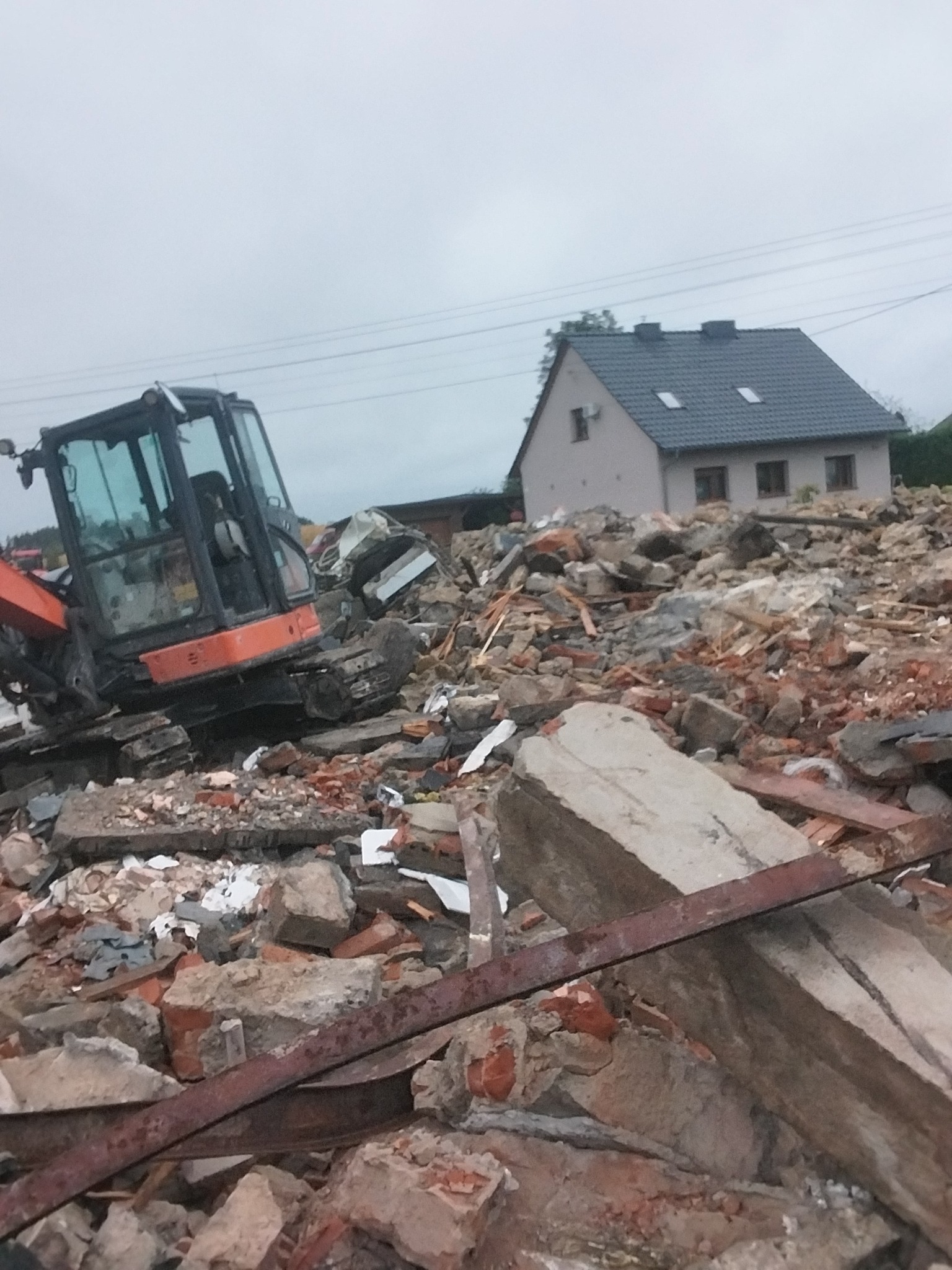 Pomarańczowa koparka na stercie gruzu po rozbiórce budynku, z widocznym fragmentem domu w tle, na pochmurnym niebie.