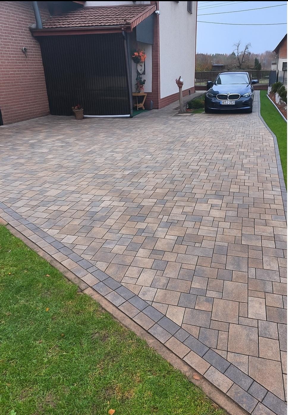 Ułożona kostka brukowa w odcieniach beżu i szarości tworzy wzór na podjeździe. Obok trawnik i zaparkowany samochód. Widoczny fragment domu z garażem.