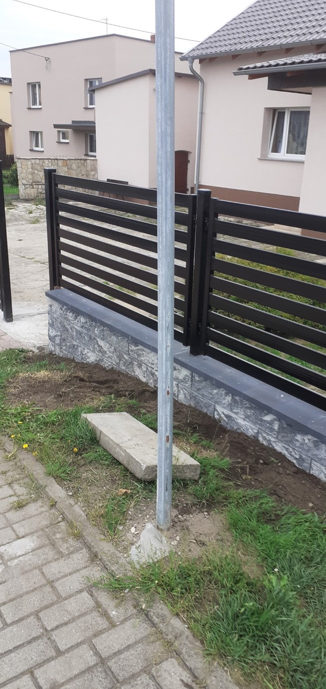 Nowoczesne ogrodzenie panelowe z poziomymi przęsłami w kolorze czarnym, osadzone na podmurówce z szarej kostki. Widok z poziomu gruntu, trawa i kostka brukowa w tle.
