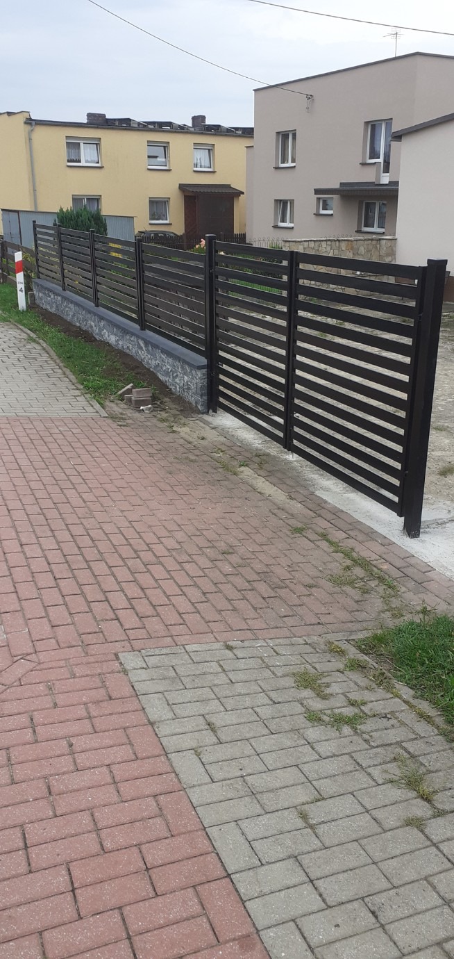Nowoczesne ogrodzenie panelowe z poziomych, czarnych listew, osadzone na niskim, kamiennym murku. Widok z chodnika z kostki brukowej, w tle budynki mieszkalne.