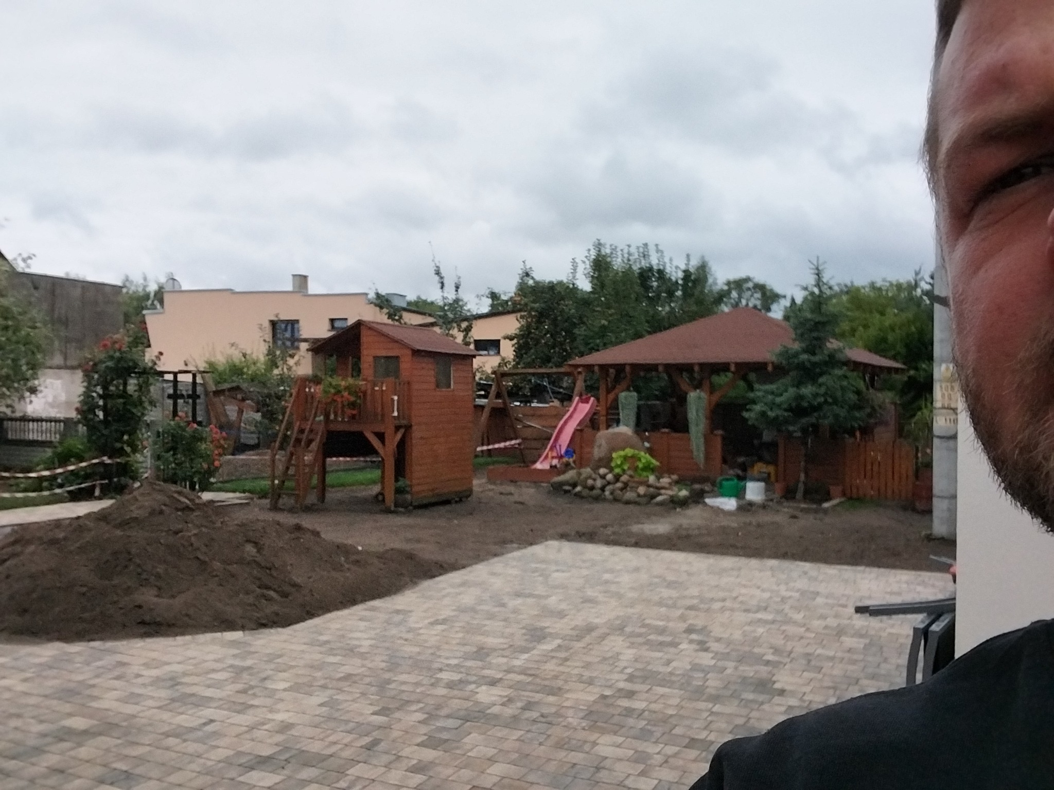 Ułożona kostka brukowa na posesji z placem zabaw w tle, widok z perspektywy osoby z boku kadru. Świeżo usypana ziemia czeka na zagospodarowanie.