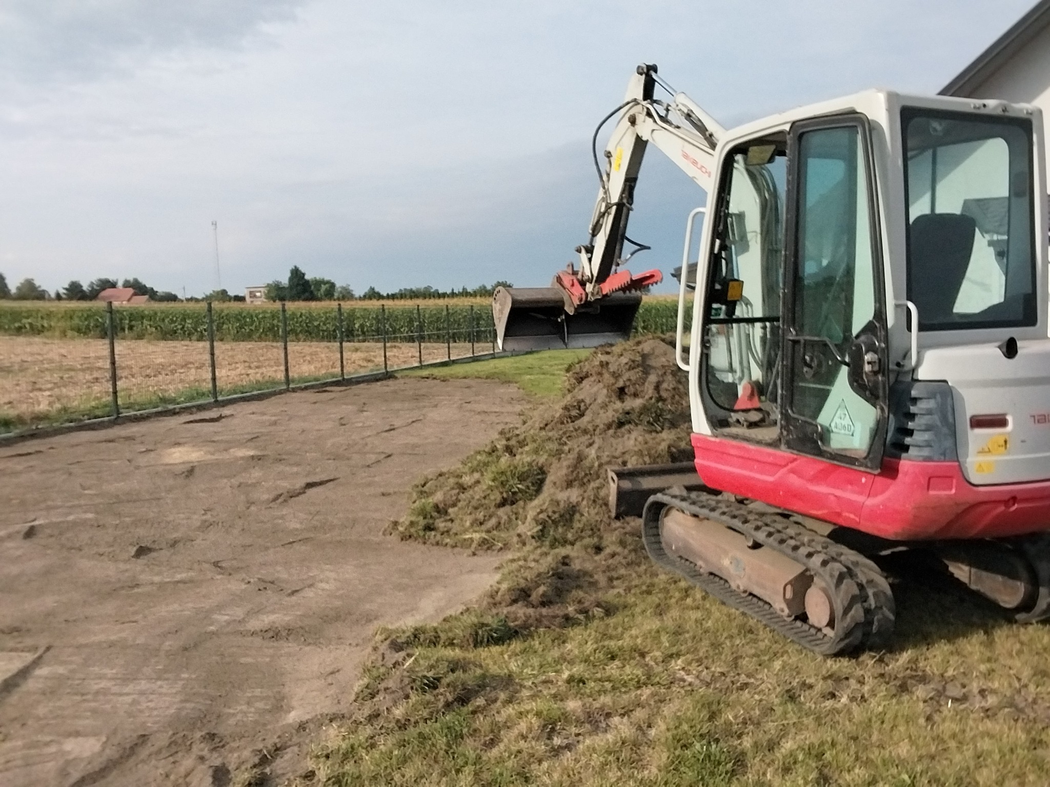 Biało-czerwona koparka gąsienicowa na polu z wykopaną ziemią, w tle ogrodzenie panelowe i uprawne pole pod pochmurnym niebem. Prace ziemne w toku.
