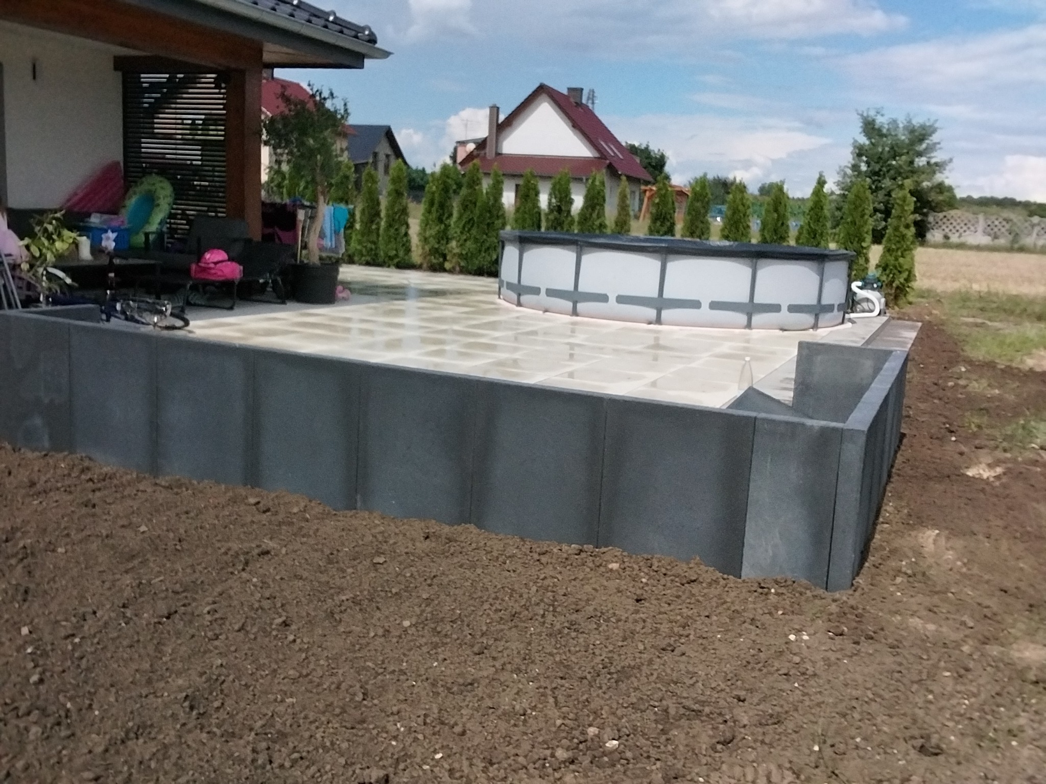 Wykończony taras z kostki przy basenie ogrodowym, otoczony niskim murem z szarych bloczków. W tle dom z ogrodem i drzewami.