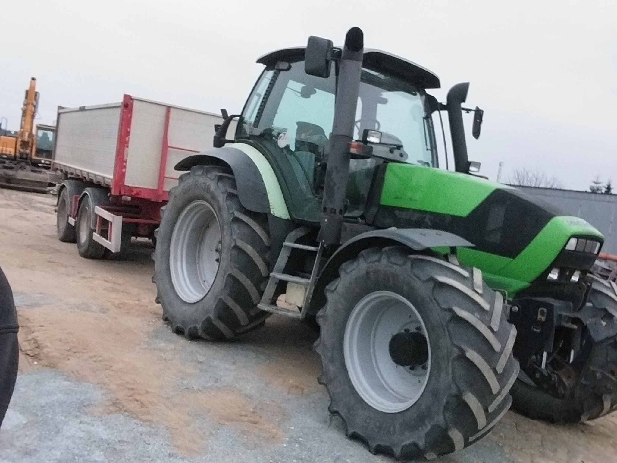 Zielony traktor z przyczepą na placu budowy, w tle widoczna koparka. Ujęcie z boku, podkreślające masywność maszyny i jej gotowość do pracy.