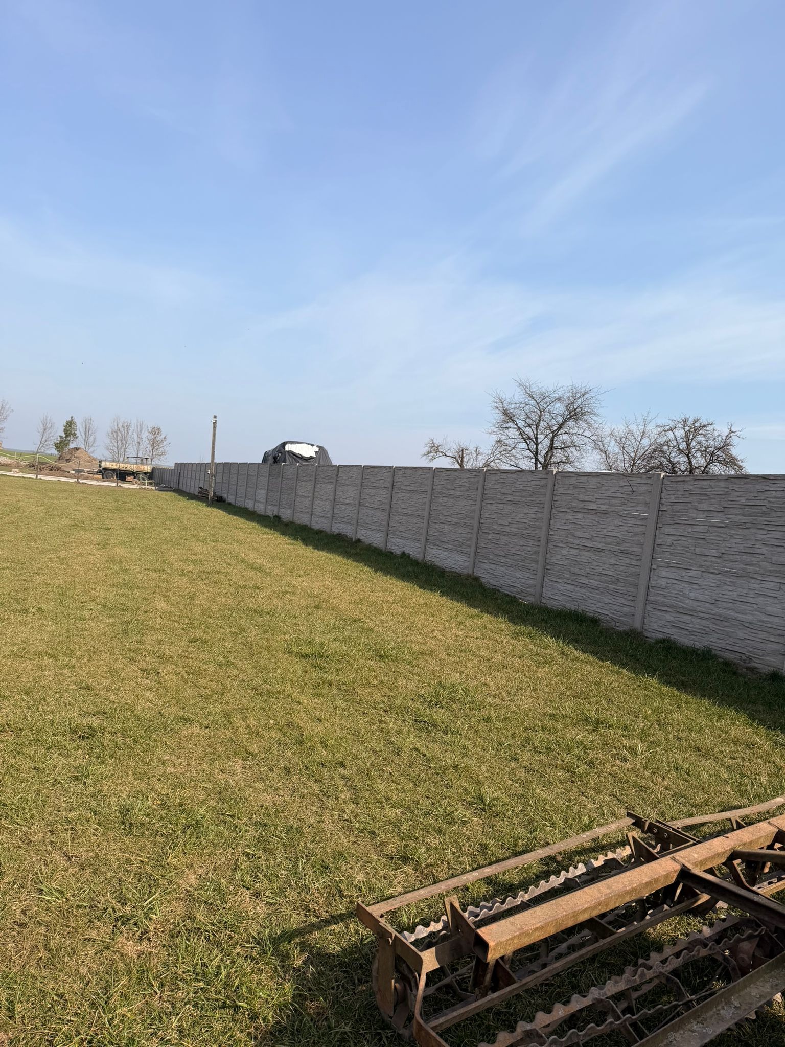 Długi, szary, betonowy płot panelowy na zielonym trawniku pod błękitnym niebem. W rogu fragment brony talerzowej. Widok na posesję.