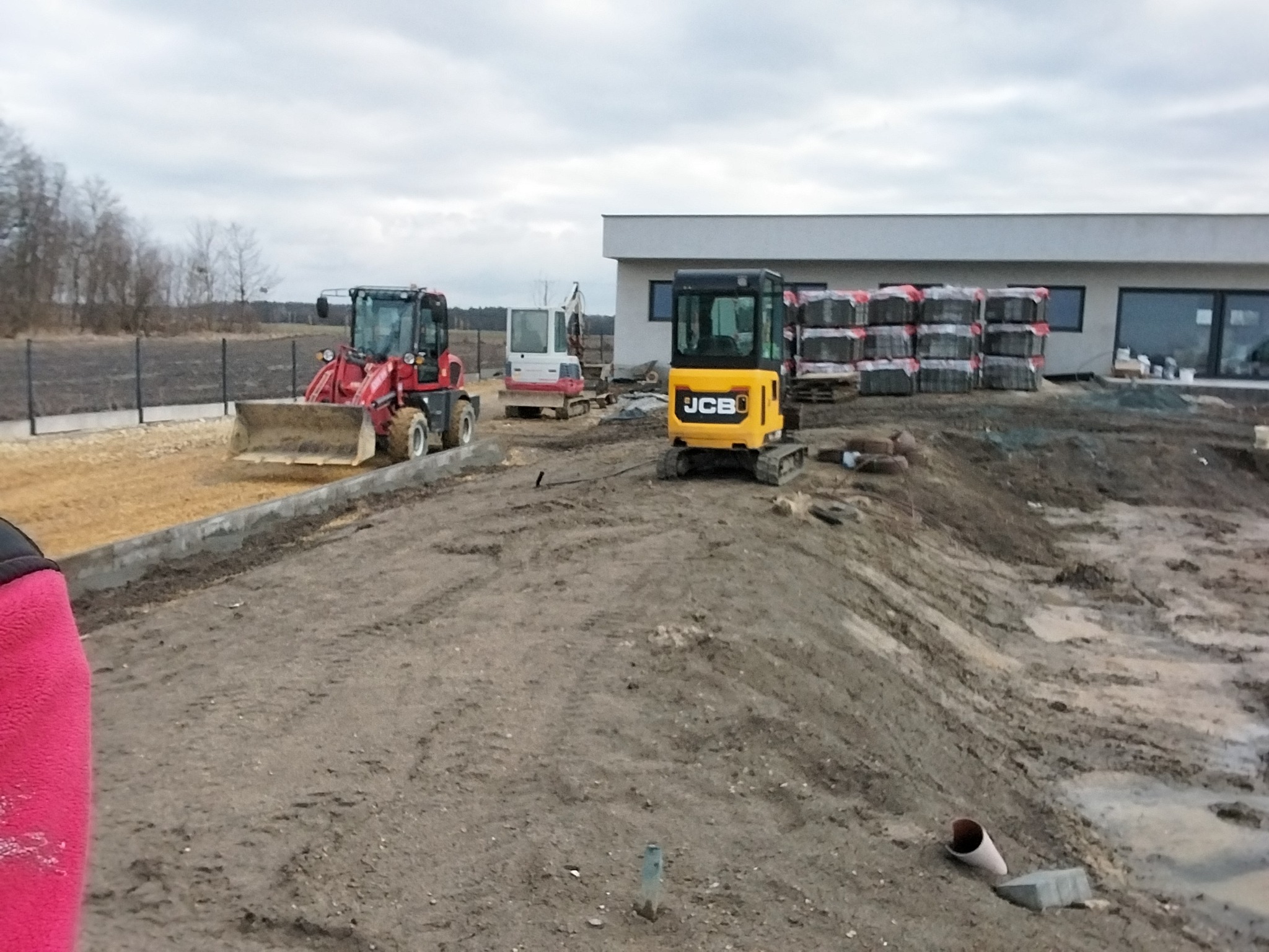 Praca ziemne: Żółta mini koparka JCB na nierównym terenie budowy, w tle czerwona ładowarka i palety z kostką brukową przy budynku. Pochmurne niebo.