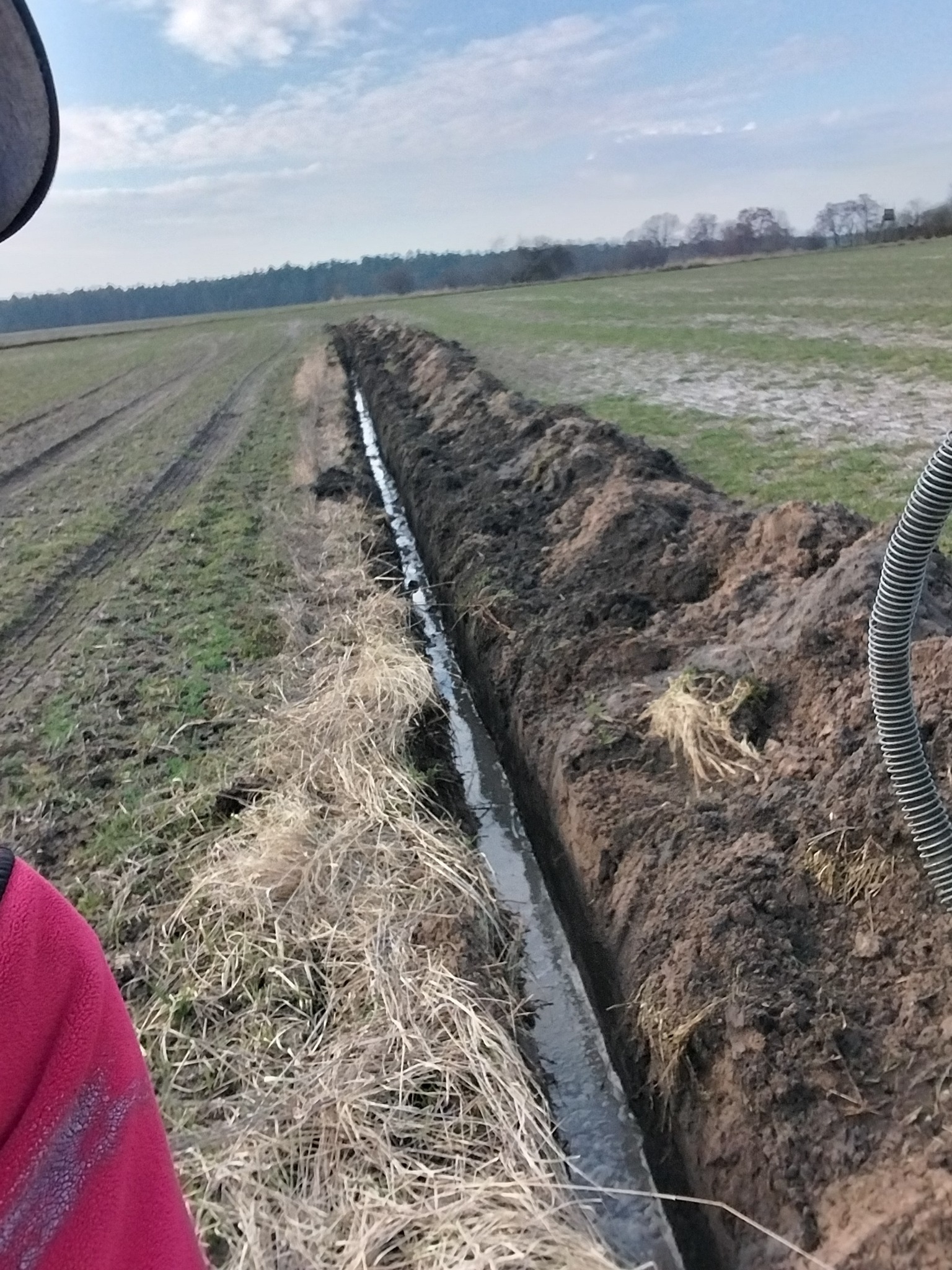 Wykopany rów melioracyjny na polu, wypełniony wodą. Widoczne ślady opon i resztki roślinności. W tle las i błękitne niebo.