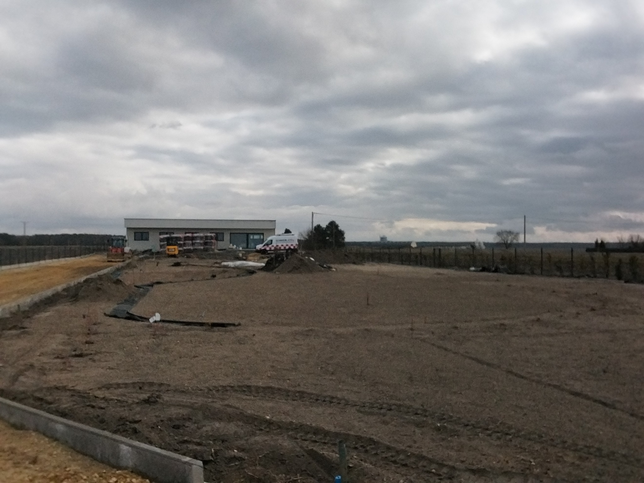Teren budowy z przygotowanym podłożem, widoczne maszyny budowlane i budynek w tle pod pochmurnym niebem. Ślady opon na ziemi.