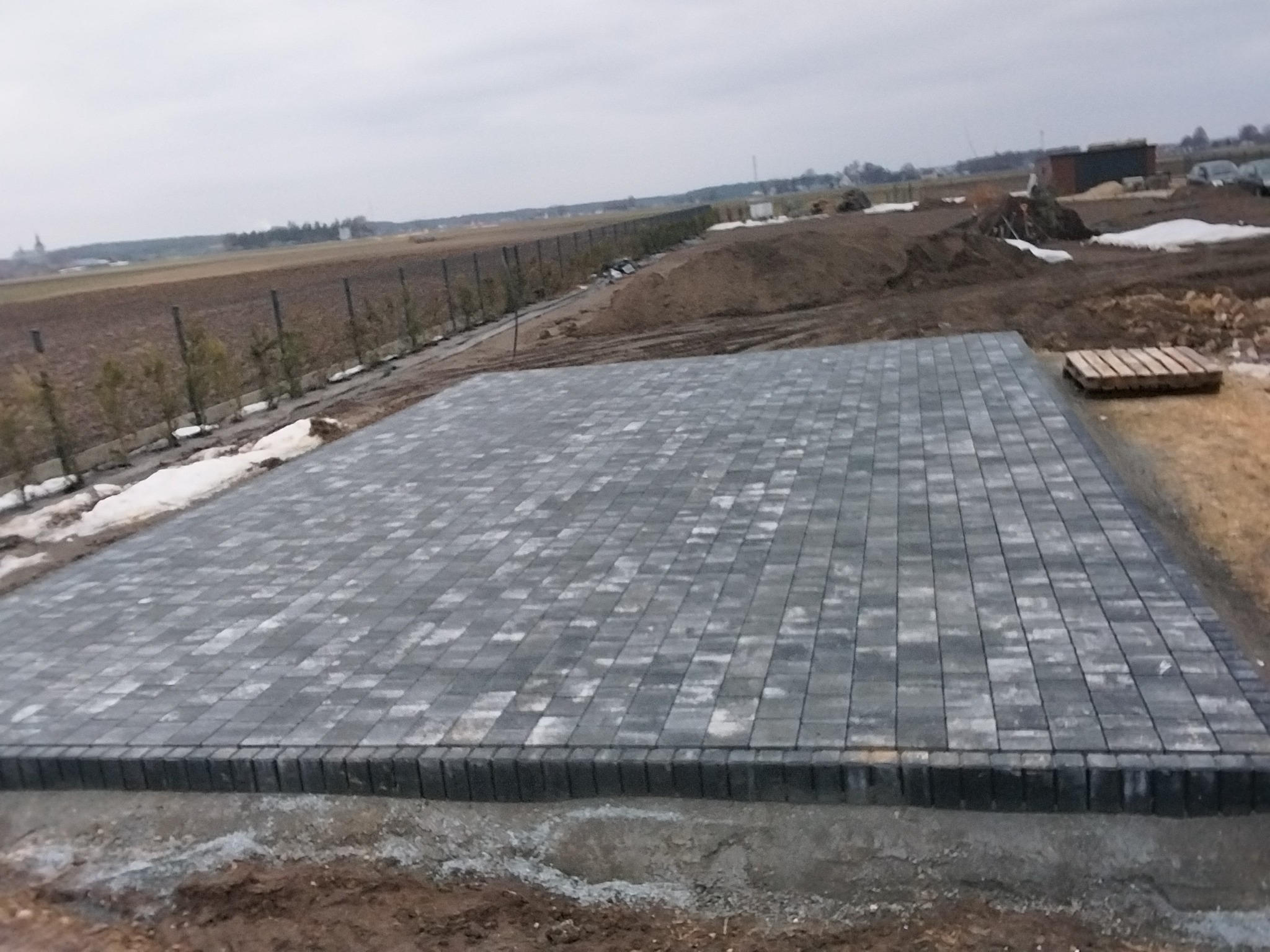 Ułożona, szara kostka brukowa na przygotowanym podłożu. Widoczne obrzeża, w tle krajobraz z drzewami i polem. Częściowo leży śnieg.