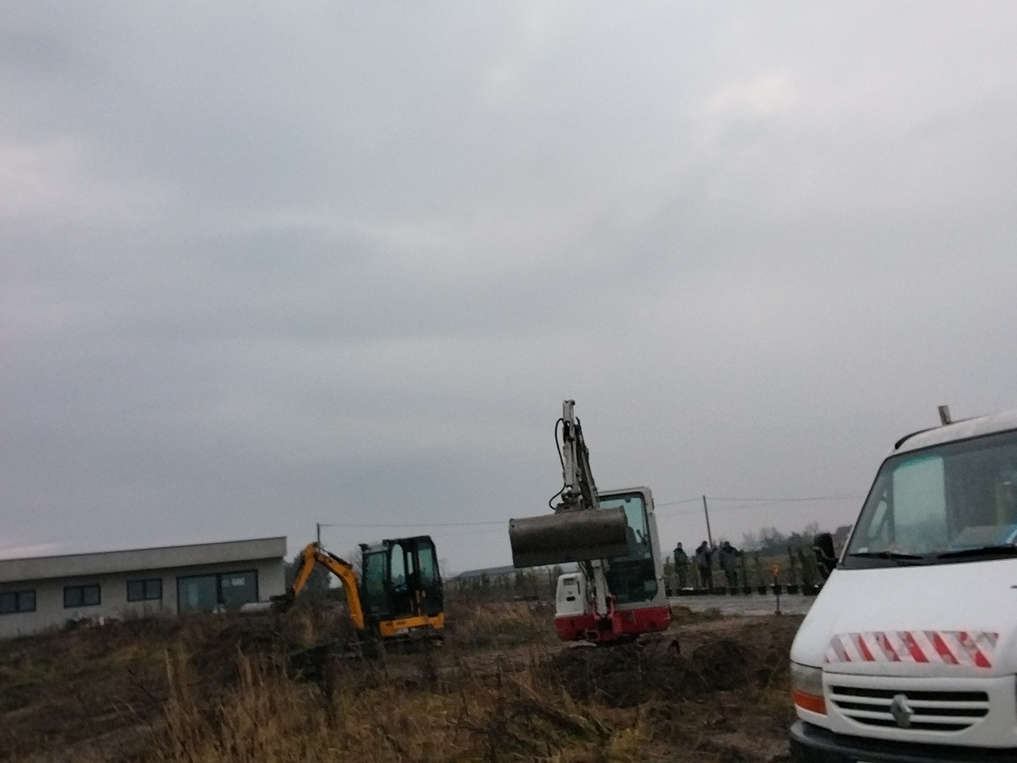 Dwie koparki na placu budowy, jedna z podniesioną łyżką, w tle budynek i grupa osób, z prawej strony kadru fragment białego samochodu dostawczego.