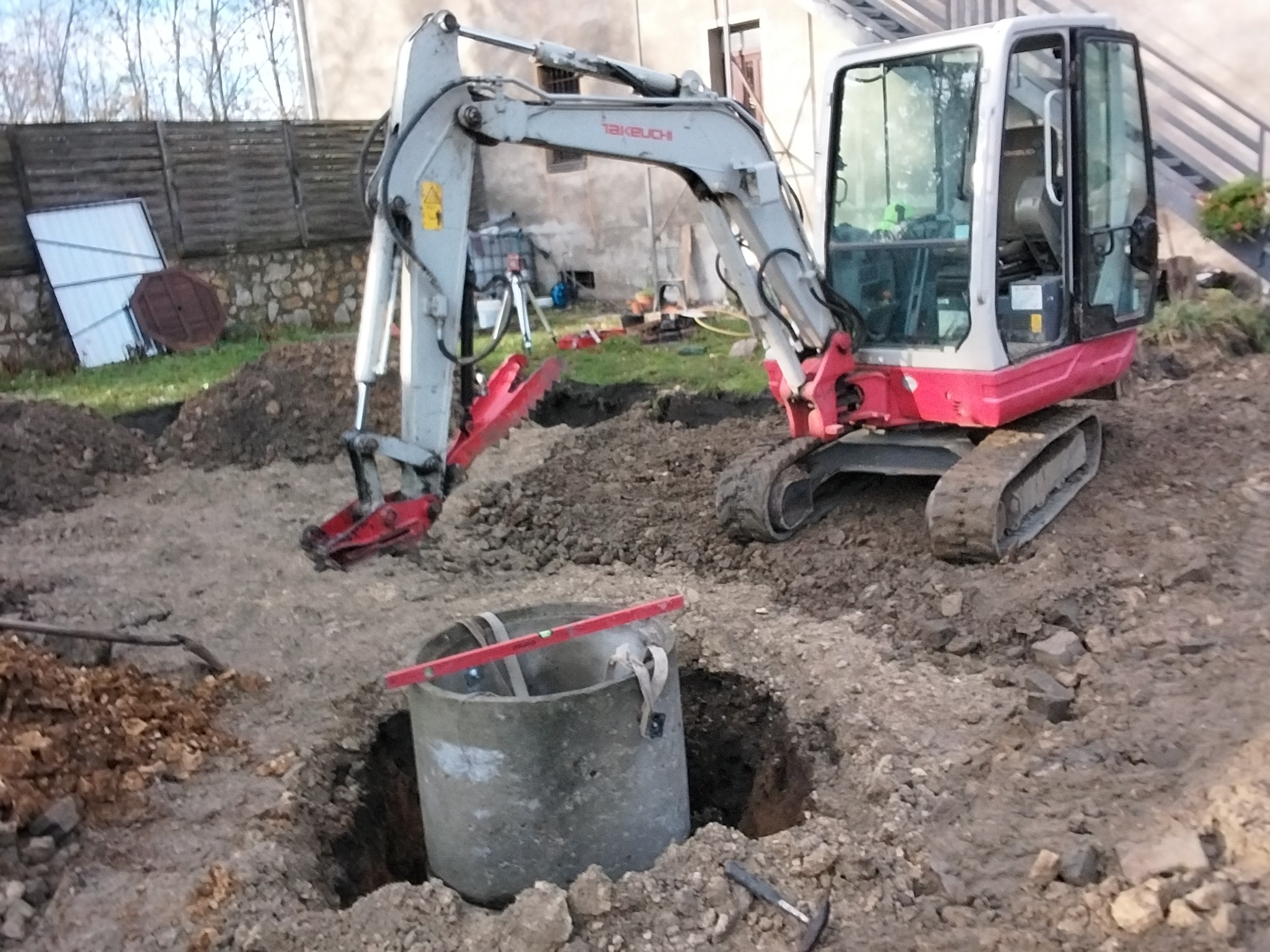 Mini koparka Takeuchi przy betonowym kręgu w wykopie. W tle budynek, ogrodzenie i drzewa. Widoczne narzędzia i poziomica. Prace ziemne w toku.