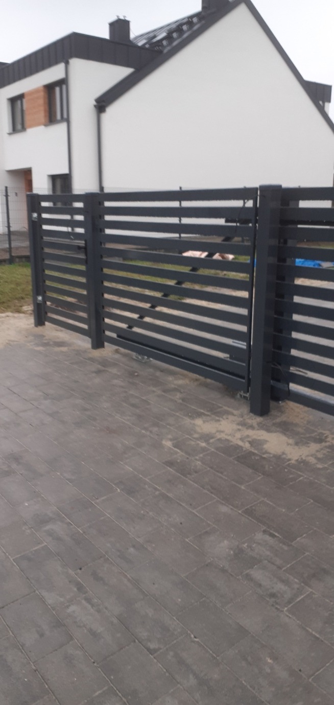 Nowoczesne, poziome ogrodzenie panelowe w kolorze grafitowym, z widoczną kostką brukową i fragmentem białego domu w tle. Minimalistyczny design, świeżo po montażu.