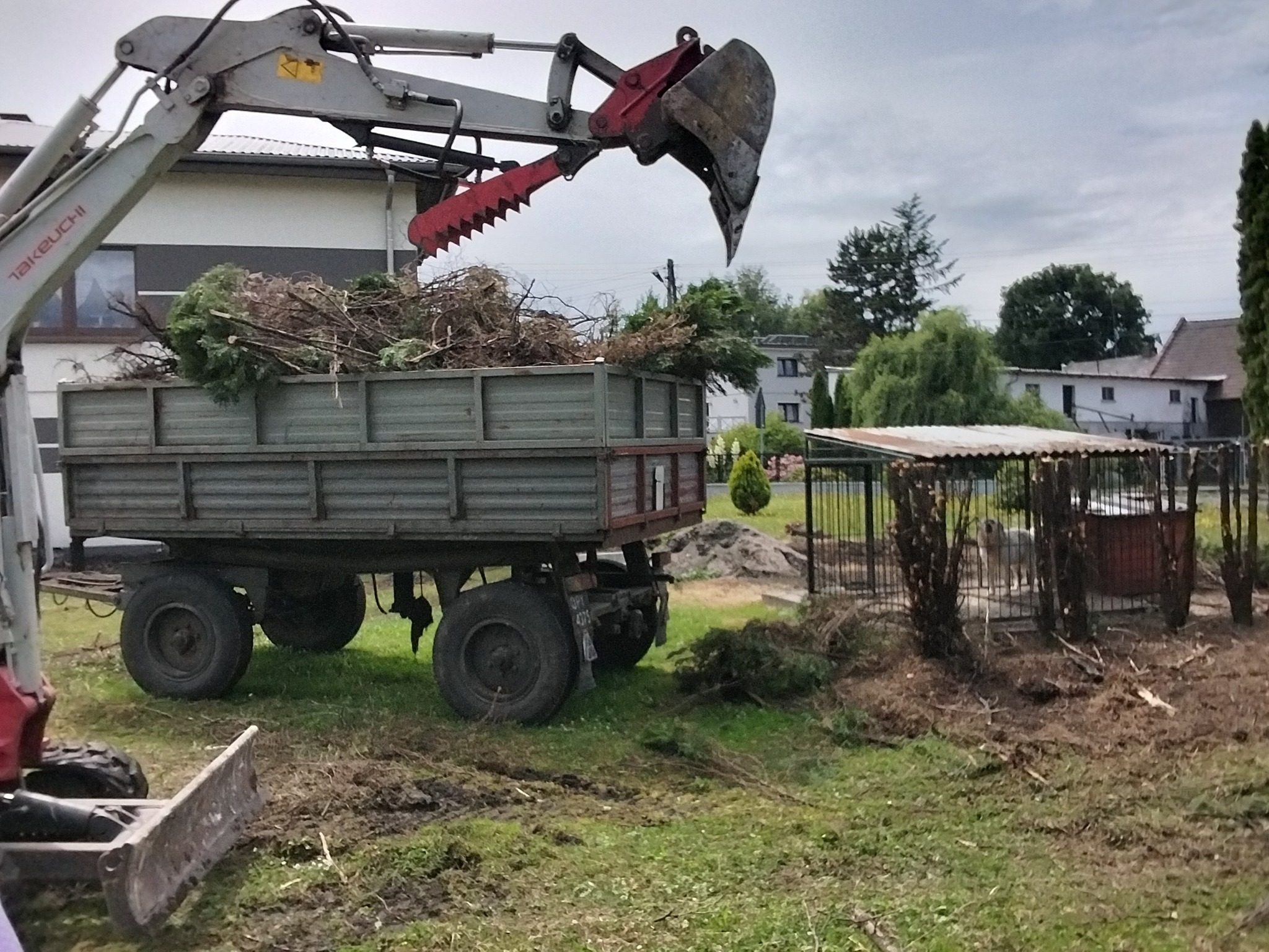 Koparka z chwytakiem załadowuje gałęzie do przyczepy na tle zabudowań. Widoczne ślady pracy w terenie.