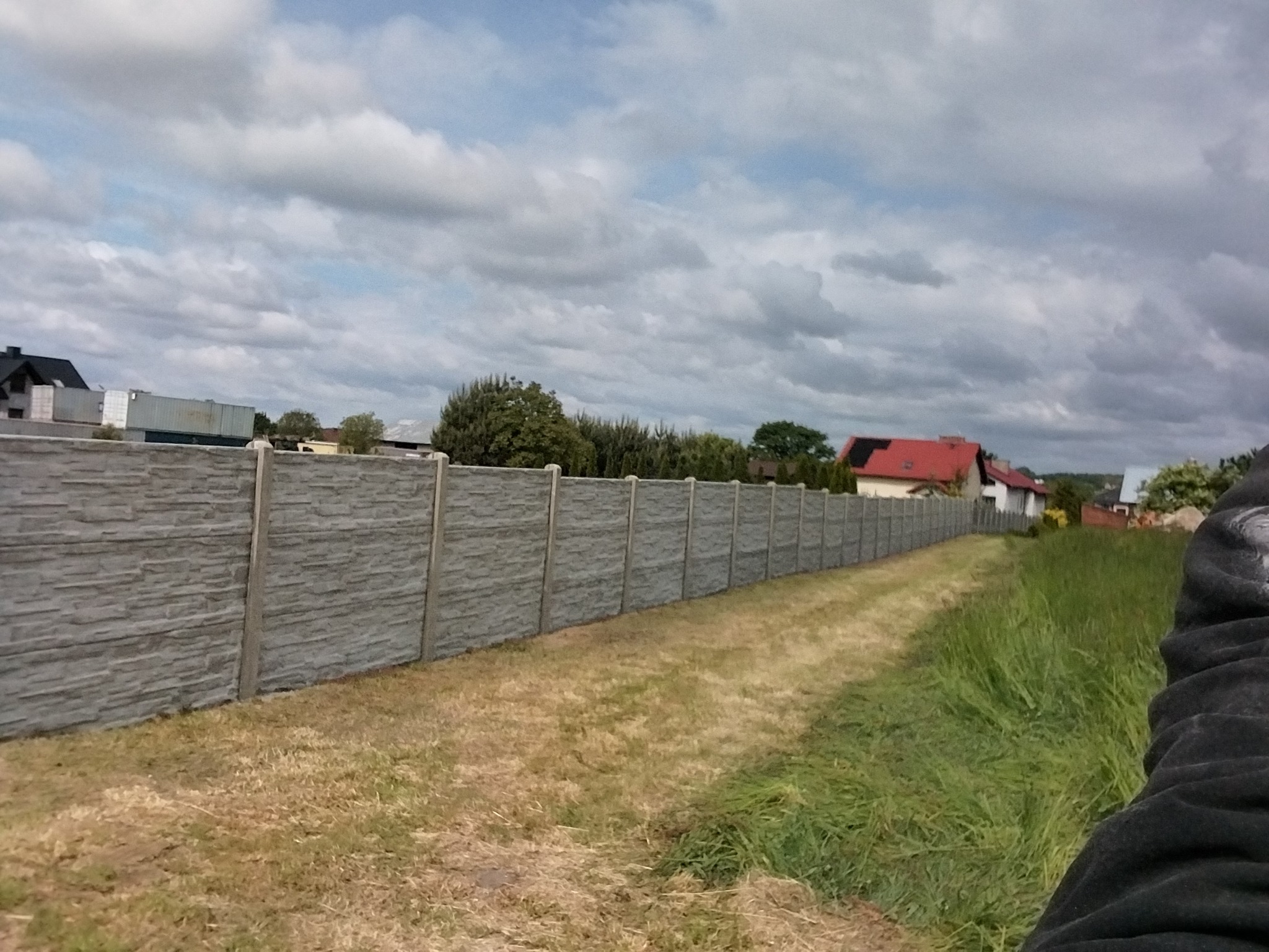 Długi, szary, betonowy płot panelowy oddzielający posesje, widziany z perspektywy trawnika. W tle domy i pochmurne niebo.